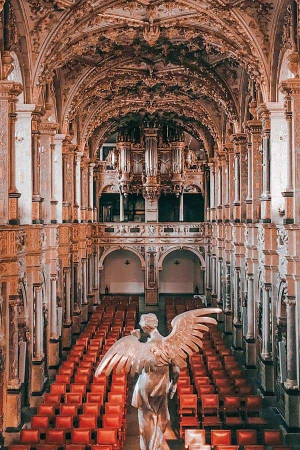 The ornate Baroque interior of Frederiksborg Castle Chapel in Hillerød, with red velvet seats, carved columns, and a winged angel statue in the foreground