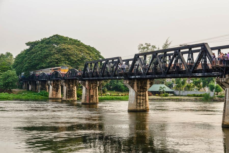 a historical black steel bridge known as the river kwai bridge that's part of the death railway in kanchanaburi near bangkok in thailand with a train crossing and visitors on the pedestrian path surrounded by lush greens and a river.