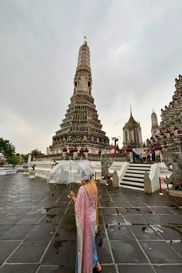 a girl in a traditional thai costume holding a clear umbrella stands facing the ornate spire or prang of wat arun, the temple of dawn on a cloudy day in bangkok, thailand