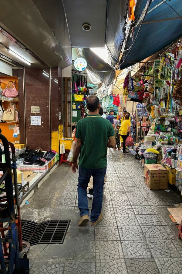 a bustling market scene in chinatown, bangkok, with a person walking down the narrow sampeng lane surrounded by shops selling colorful goods like bags, accessories, and household items