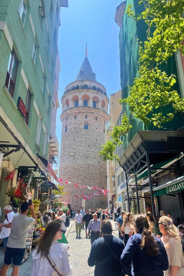a bustling street in Istanbul lined with shops and cafes, leads to the historic galata tower and numerous people are walking and taking photos with the sun shining brightly on this lively scene