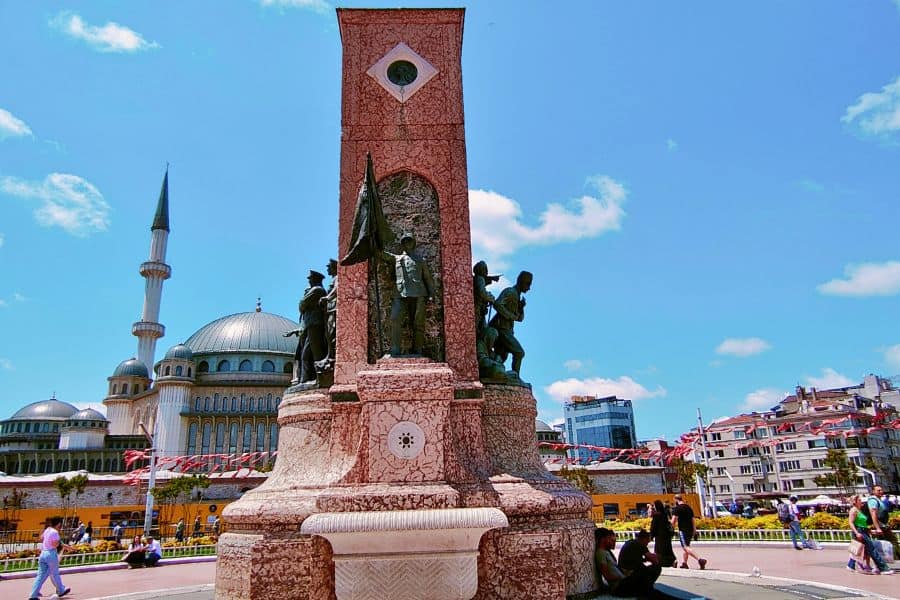 statue with a mosque in the background under a bright blue sky in taksim square in Istanbul where people are walking and sitting