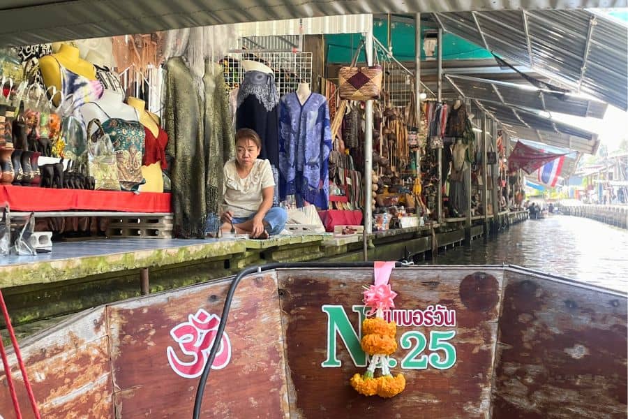 view from a boat of a vendor sitting by a colorful clothing stall at damnoen saduak floating market in bangkok