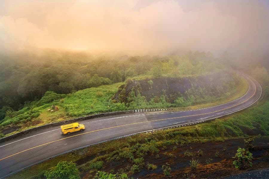winding mountain road surrounded by lush greenery and mist, with a yellow vehicle driving through the scenic landscape