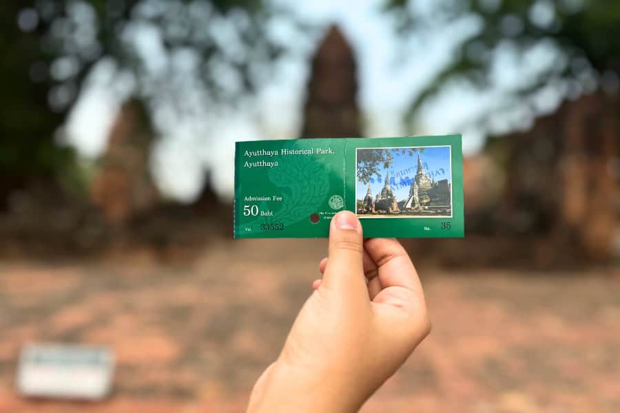close-up of mini-me's hand holding an entrance ticket to ayutthaya historical park in thailand, with ancient temple ruins blurred in the background