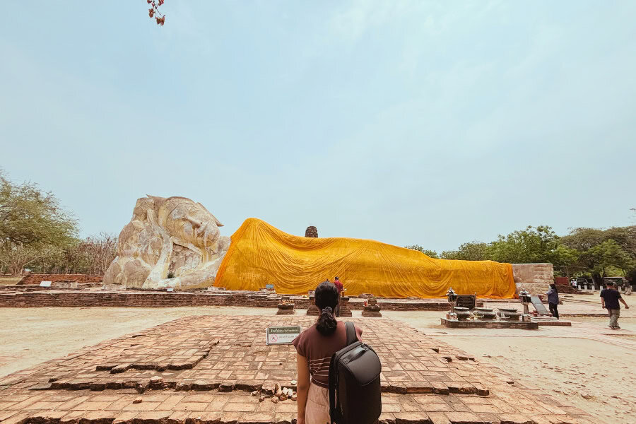 mini-me walking towards the massive reclining buddha at wat lokayasutharam in ayutthaya, thailand, draped in a bright yellow cloth, with ancient brick ruins surrounding the site