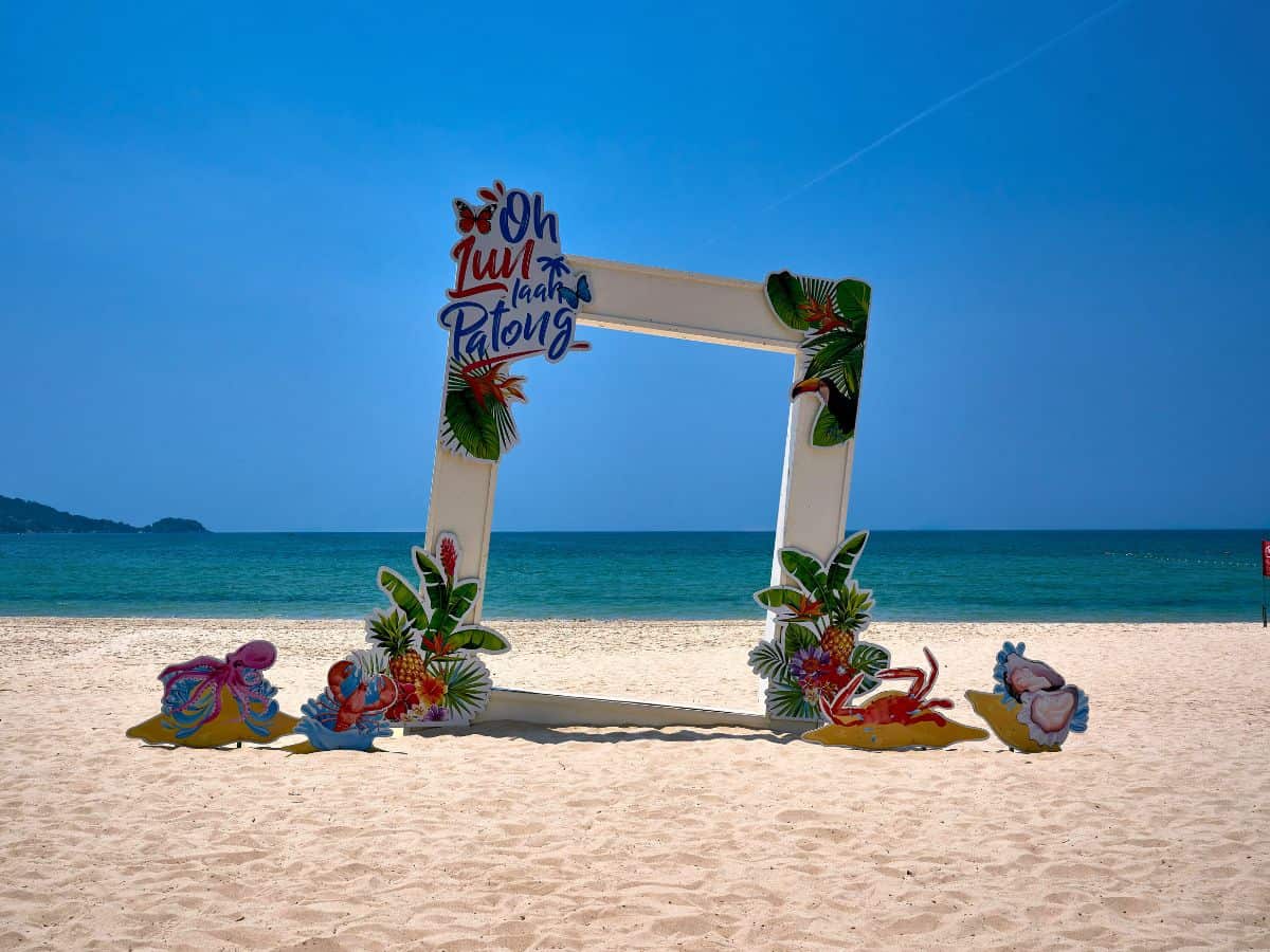 beachfront photo frame decoration with tropical elements and 'oh luu haa patong' sign on the sandy shore of patong beach, phuket, with the blue ocean in the background