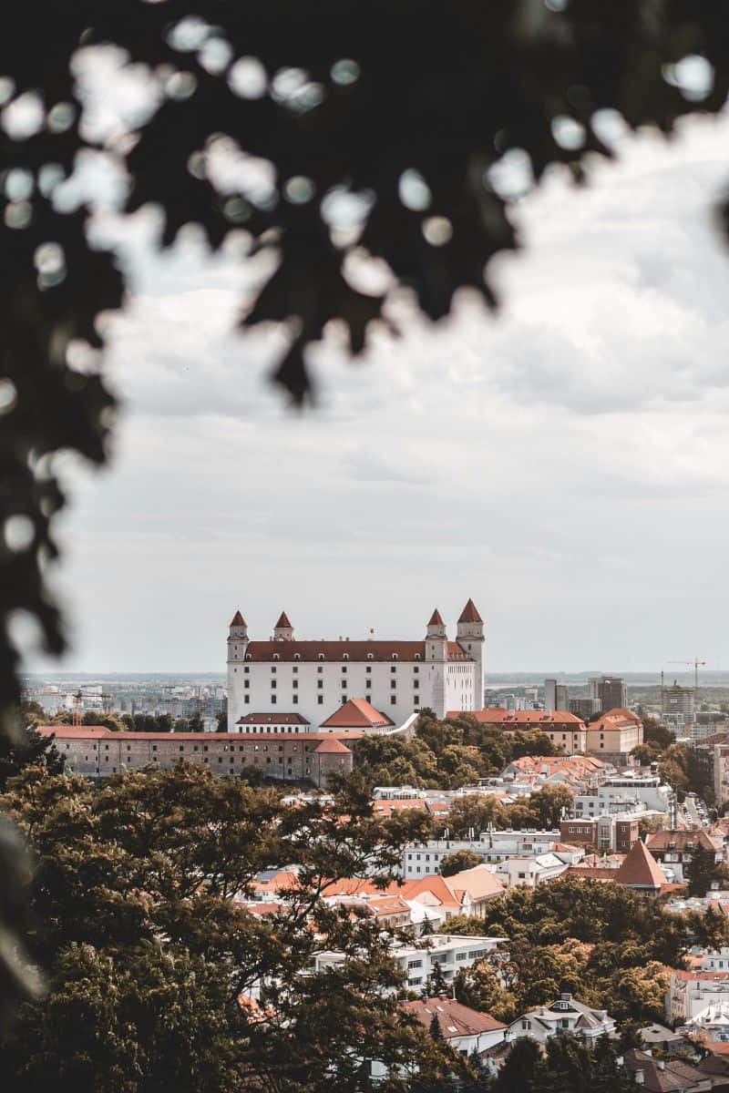 distant view of the white Bratislava Castle with its red rooftops and four corner towers, perched on a hill overlooking the city of Bratislava, Slovakia, framed by leafy branches in the foreground