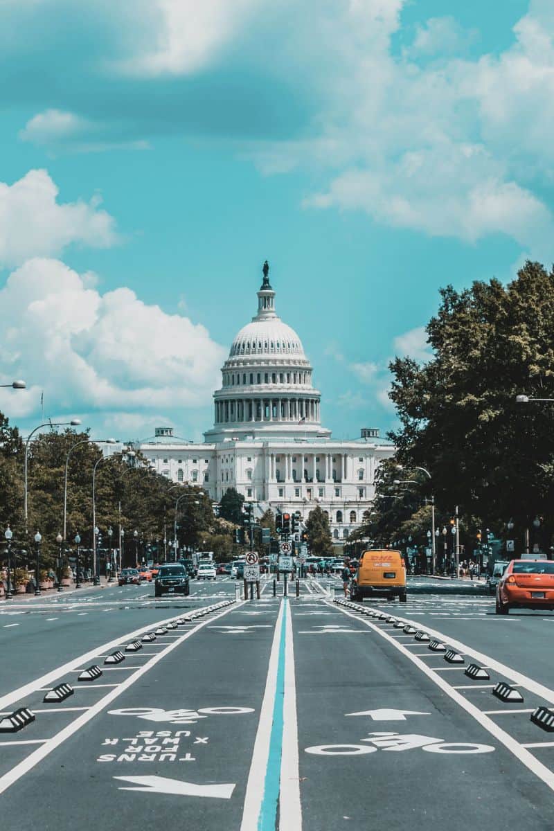united states capitol building in washington dc seen from a road lined with trees and traffic, under a bright blue sky with scattered clouds