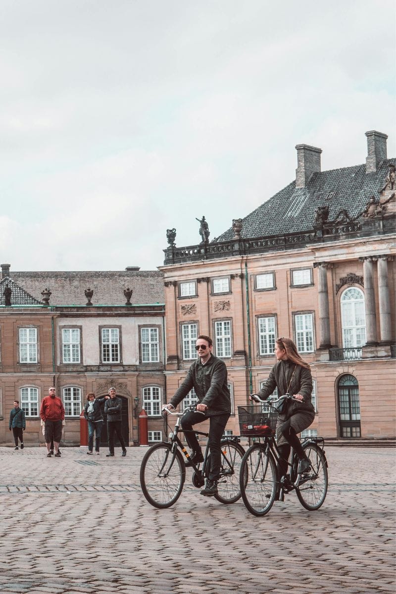 a couple riding bicycles through a cobbled square in copenhagen, denmark, with historic buildings and curious onlookers in the background
