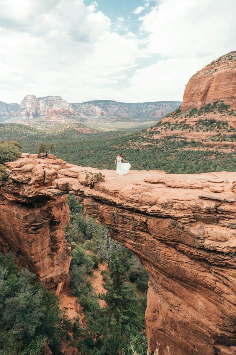 woman in a white dress standing on devil's bridge, a natural red rock formation in sedona, arizona, surrounded by dramatic cliffs and lush green forest under a partly cloudy sky