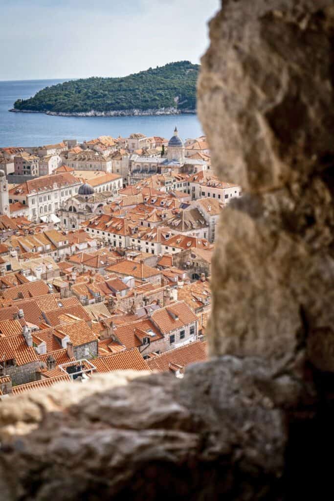 view of dubrovnik's old town with terracotta rooftops and domed churches framed through a stone window, with lokrum island and the adriatic sea in the background