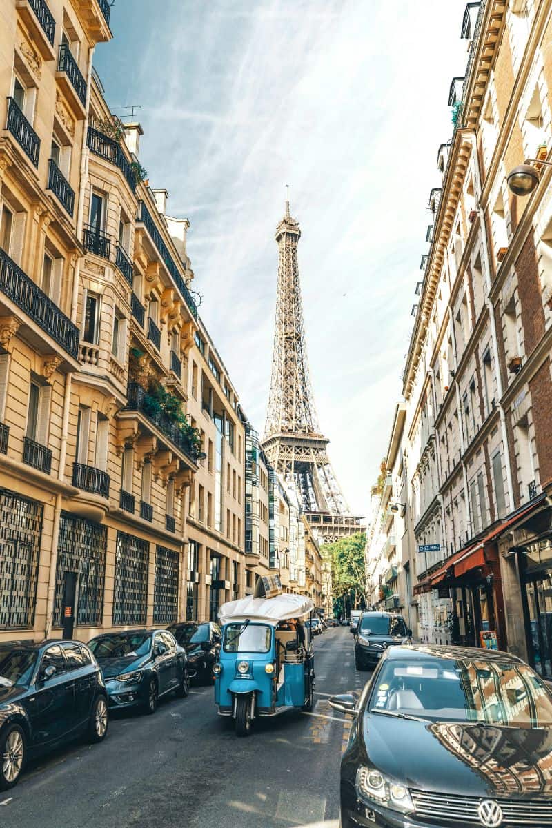 a narrow Parisian street lined with classic Haussmann-style buildings, with the Eiffel Tower rising in the distance and a blue tuk-tuk driving between parked cars