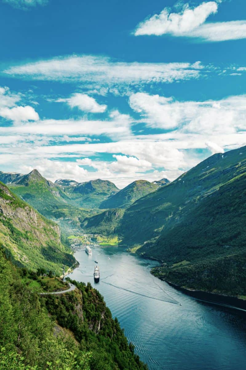  scenic view of Norway’s Geirangerfjord with cruise ships sailing through deep blue waters, surrounded by steep, green mountains and dramatic clouds under a bright blue sky