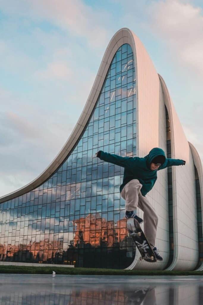 a skateboarder mid-jump in front of the futuristic heyday aliyev center in baku, azerbaijan, with its iconic curved architecture reflecting the cityscape at sunset
