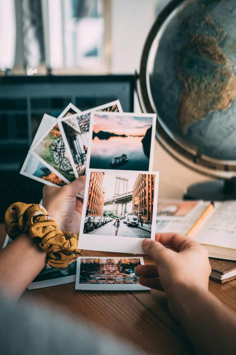hands holding printed travel photographs near a globe and travel journal on a wooden desk