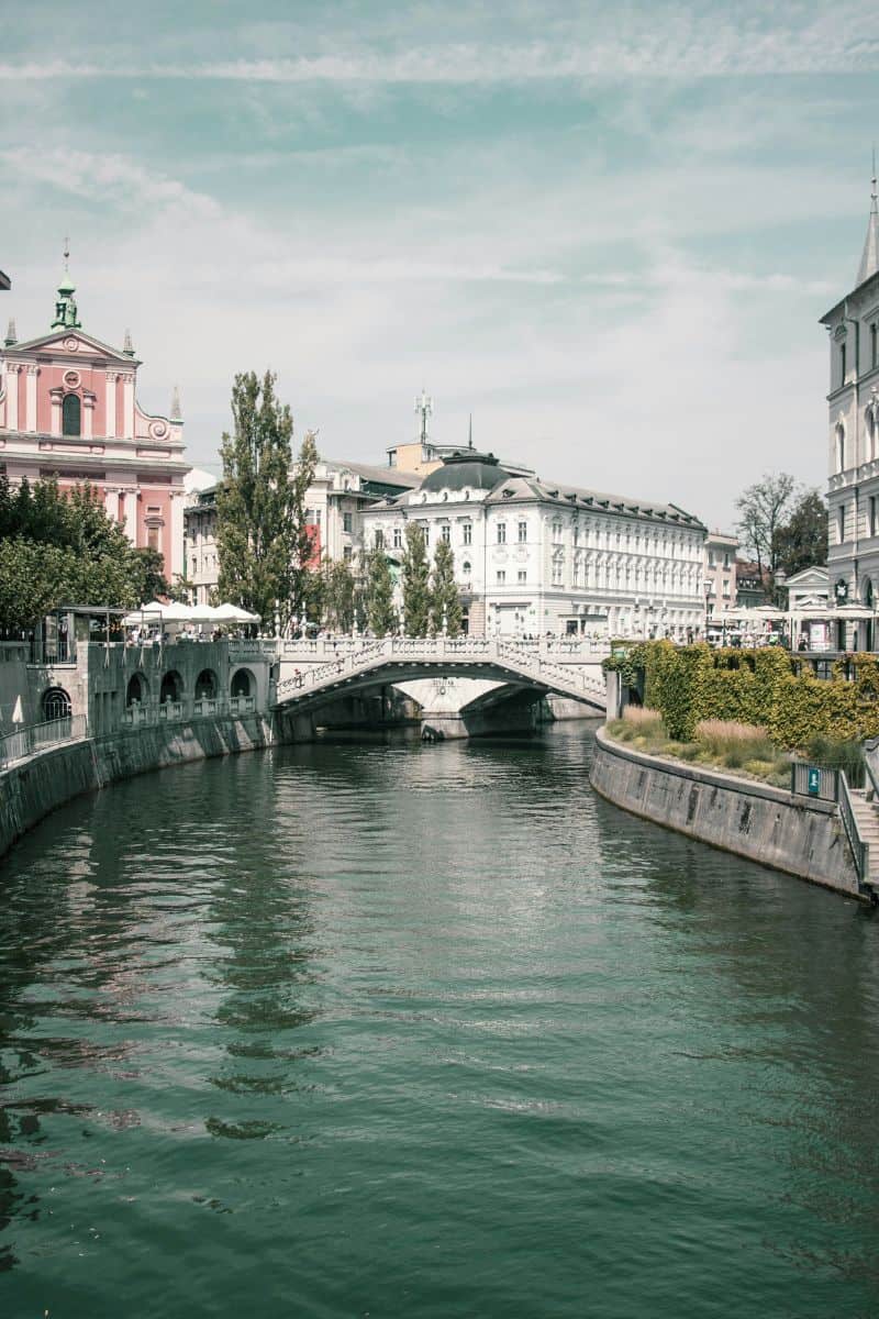 view of the Triple Bridge in Ljubljana, Slovenia, spanning the Ljubljanica River with historic white buildings and the pink Franciscan Church of the Annunciation in the background under a partly cloudy sky