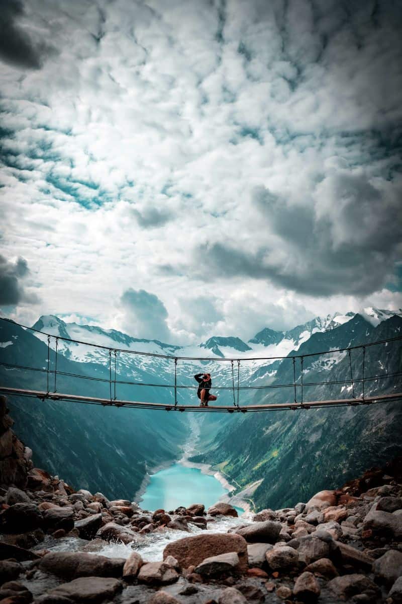 a person crouching on the iconic suspension bridge near Olpererhütte in Schmiedwiese, Mayrhofen, Austria, overlooking a turquoise lake and surrounded by the snow-capped peaks of the Austrian Alps under dramatic clouds