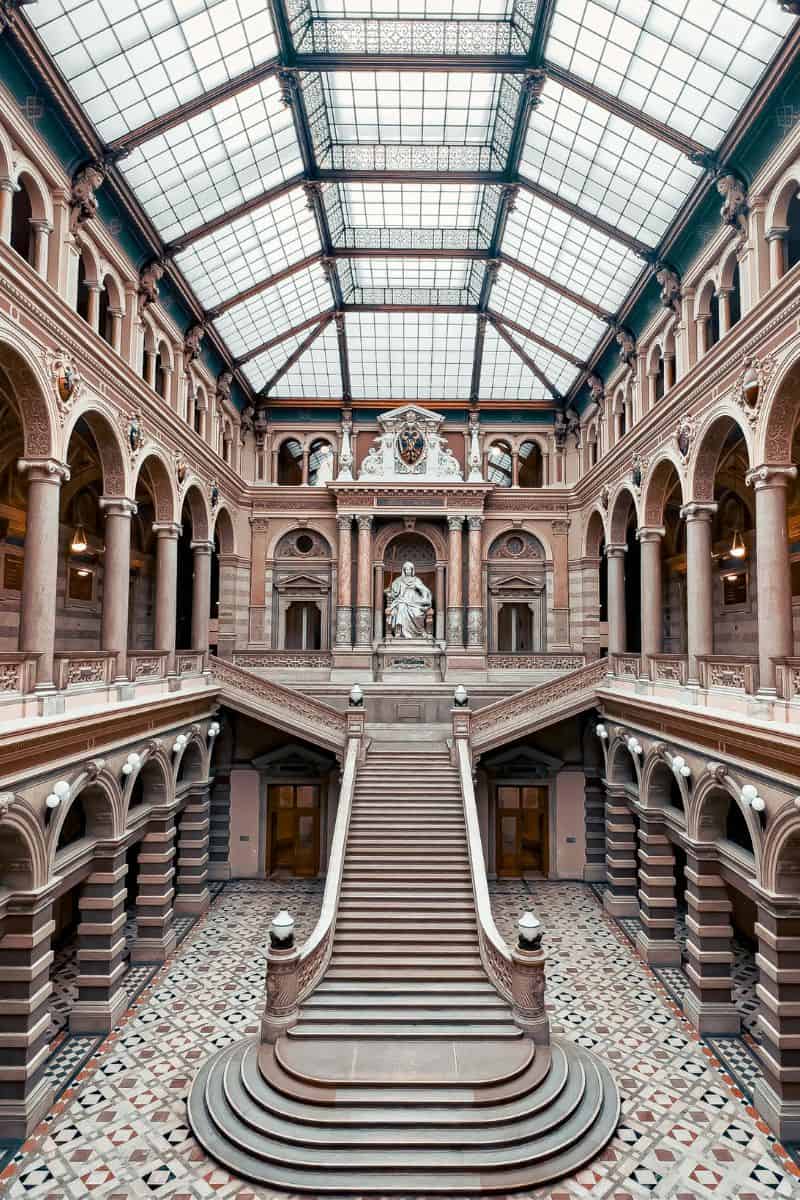 grand staircase inside the palais of justice in vienna, austria, featuring ornate arches, marble columns, a patterned floor, and a central statue beneath a striking glass ceiling
