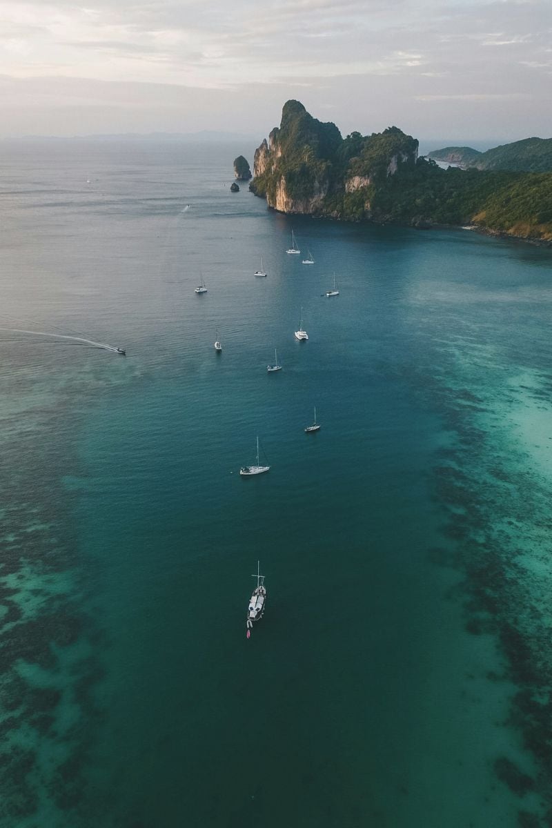 an aerial view of turquoise waters with scattered sailboats, leading towards a rocky limestone island in Thailand's Andaman Sea