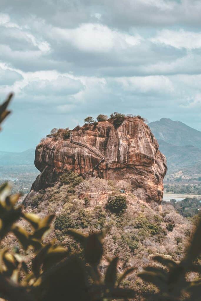  iconic Sigiriya Rock Fortress in Sri Lanka, a massive ancient rock rising from lush green surroundings with visible stairways leading to the summit, set against a backdrop of mountains and cloudy skies