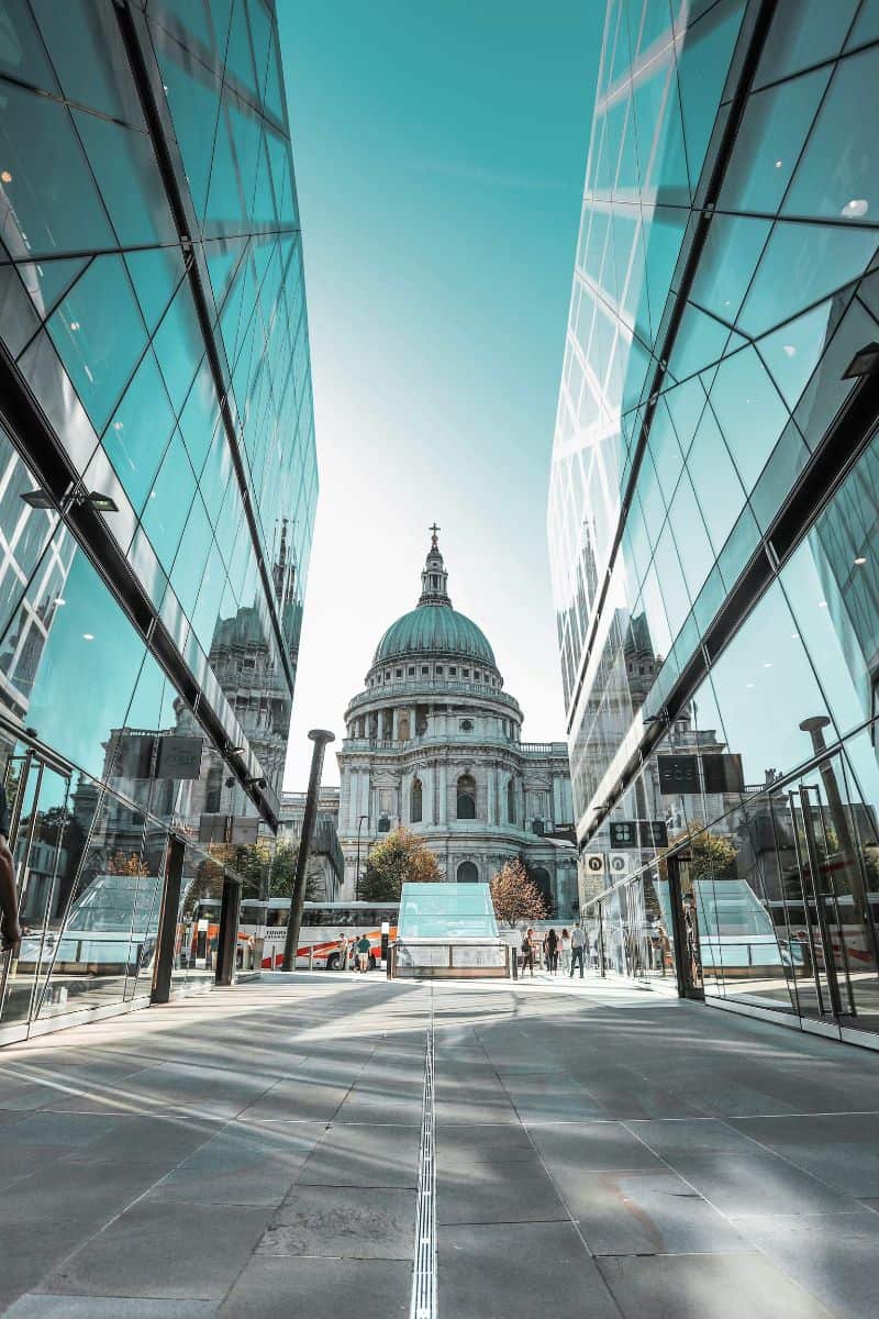 view of St. Paul’s Cathedral in London framed between two modern glass buildings, with reflections creating a symmetrical composition under a bright blue sky