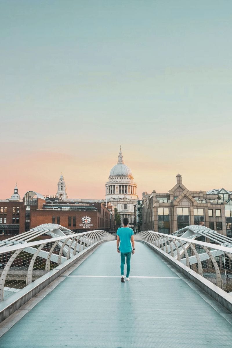 a person walking alone on the Millennium Bridge in London at sunrise, with a clear view of St. Paul's Cathedral framed by modern and historic buildings under a soft pastel sky