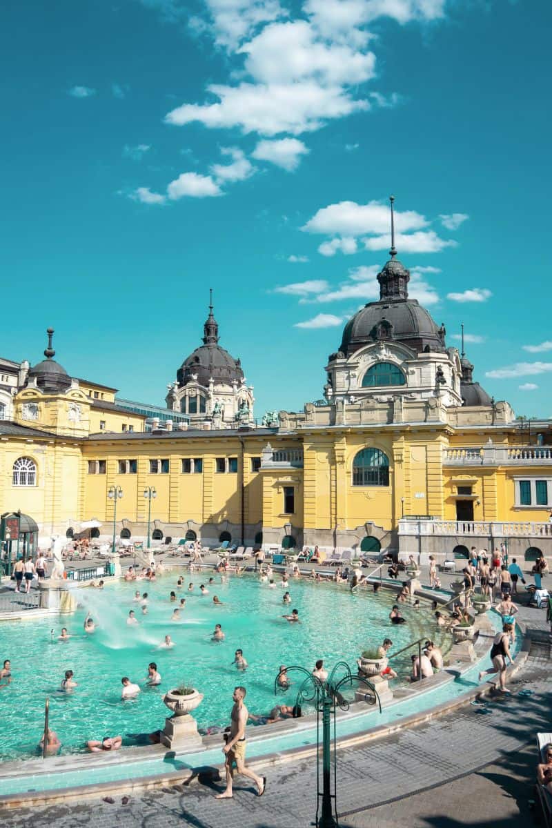 people relaxing and swimming in the iconic outdoor thermal pool at Széchenyi Thermal Bath in Budapest, Hungary, with its grand yellow neo-baroque architecture and domed towers under a bright blue sky
