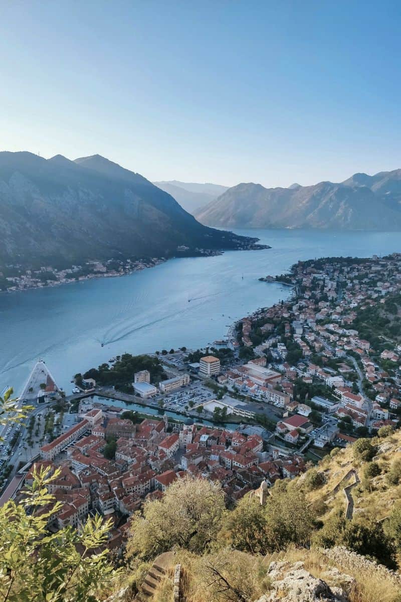  aerial view of the Bay of Kotor in Montenegro, showcasing the red-roofed old town of Kotor, surrounded by dramatic mountains and calm blue waters, with winding roads and boats visible in the bay