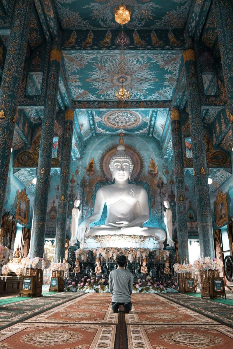 a person kneeling in prayer inside the Blue Temple (Wat Rong Suea Ten) in Chiang Rai, Thailand, with a striking white Buddha statue and ornate blue and gold interiors