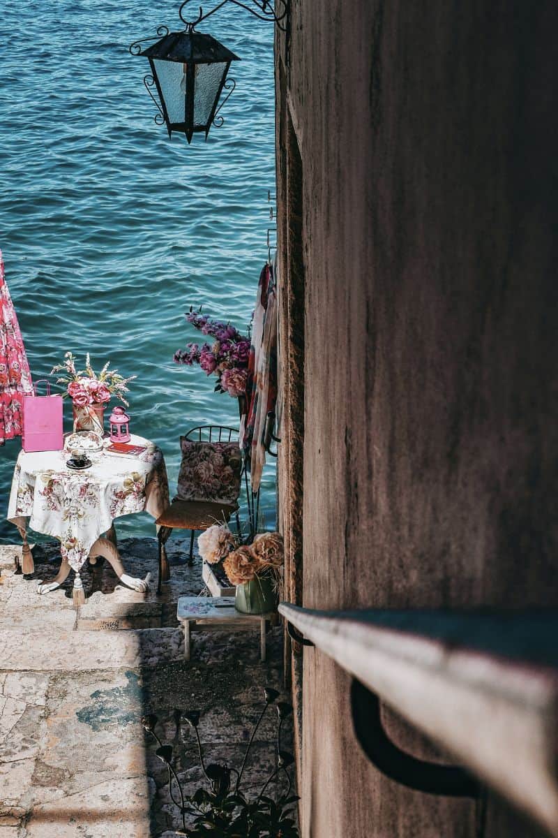 a charming waterfront café corner in Rovinj, Croatia, featuring a floral tablecloth, tea set, and decorative pink details, set against a stone wall overlooking the calm blue Adriatic Sea