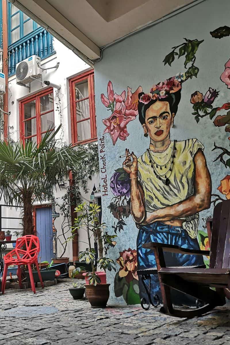 A quirky courtyard mural of Frida Kahlo at Hotel Check Point in Tbilisi, surrounded by potted plants, red chairs, and pops of color