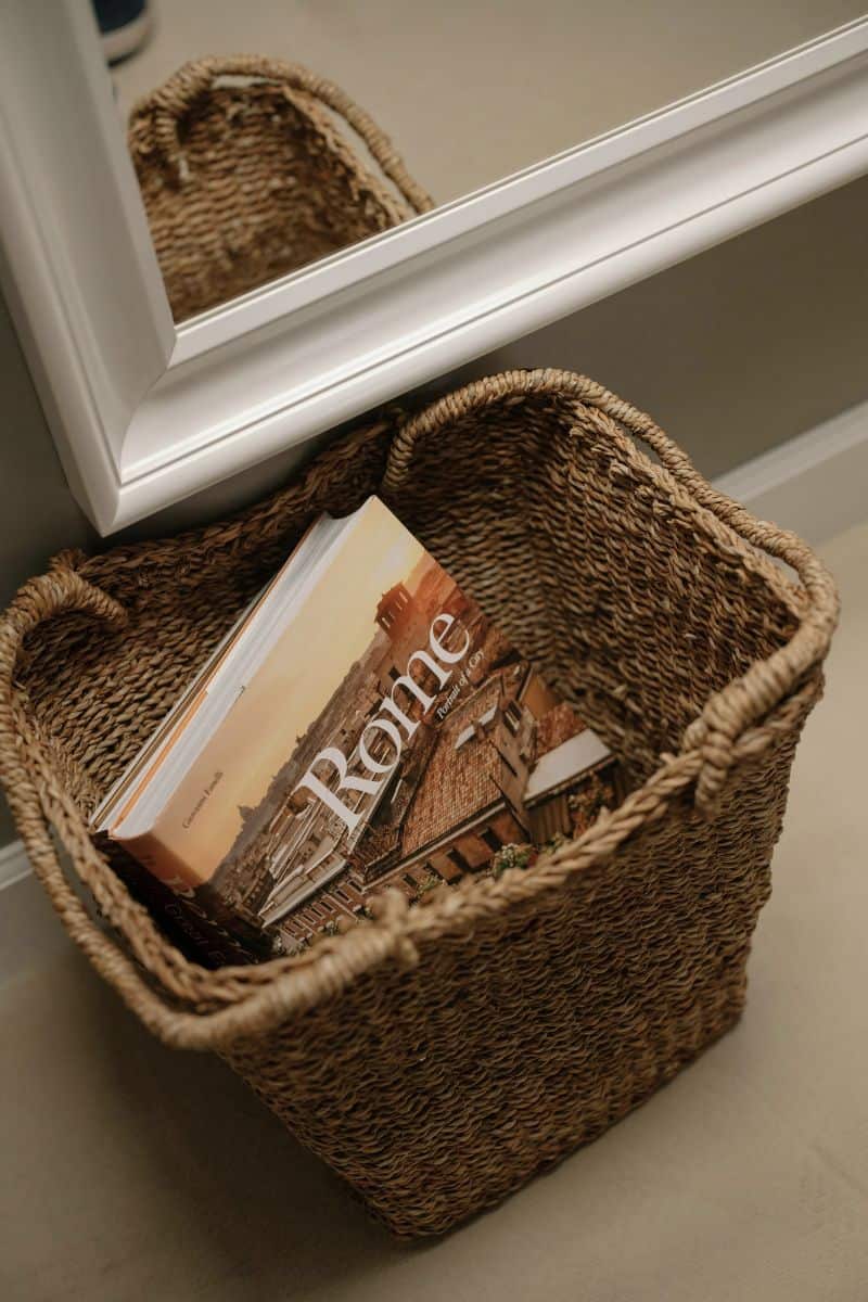 woven basket with a rome travel book placed on the floor near a white framed mirror