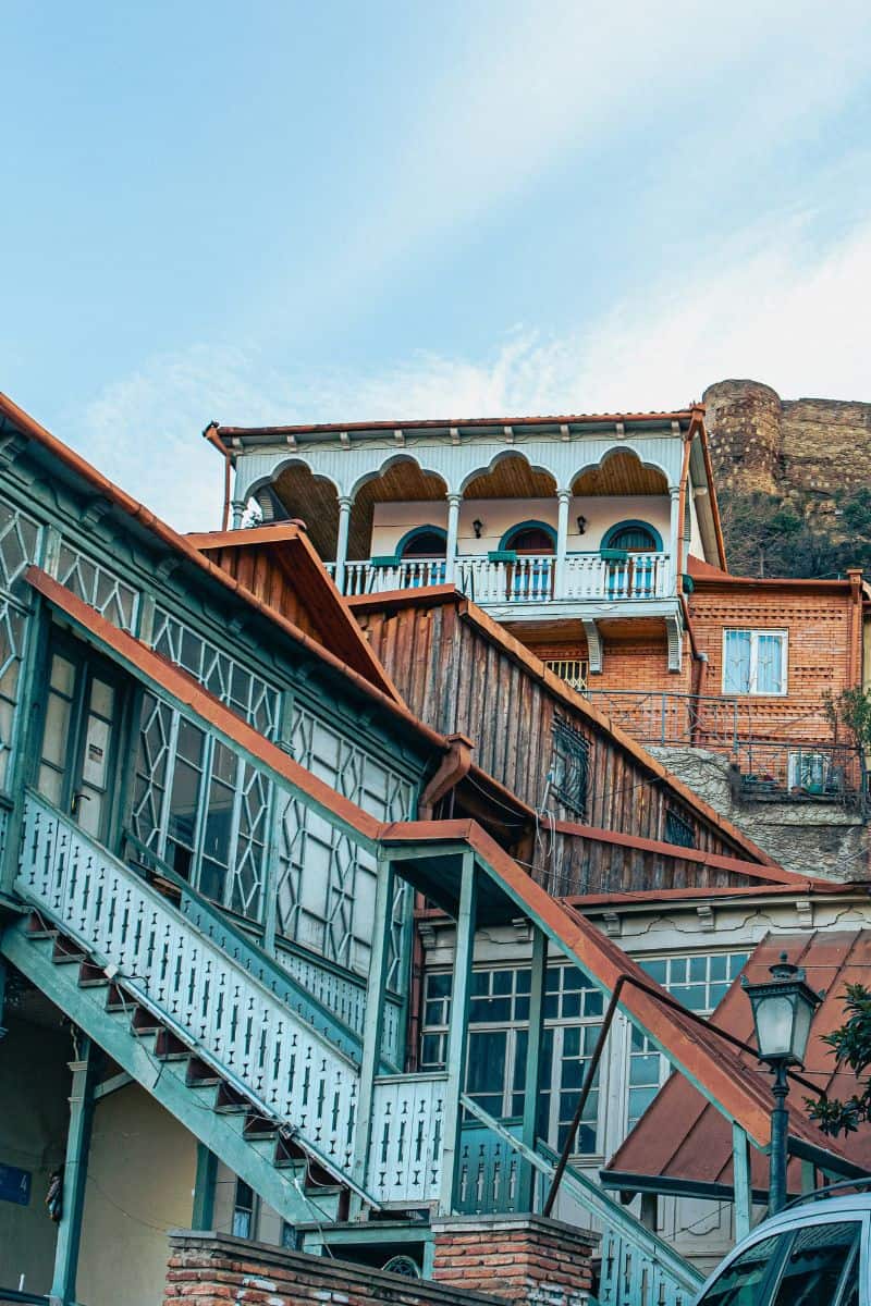 Traditional wooden houses with detailed balconies stacked along a steep hillside under Narikala Fortress in Tbilisi