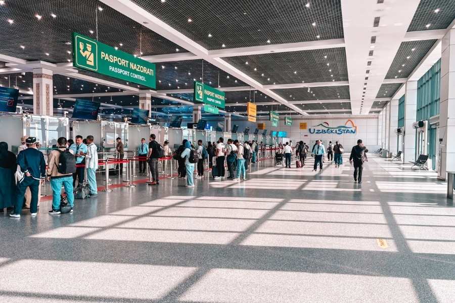 people standing in line at passport control inside tashkent international airport with signs in uzbek and english under bright sunlight