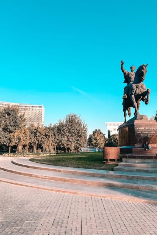 amir temur statue in tashkent, uzbekistan, showing the iconic equestrian monument with hotel uzbekistan in the background, surrounded by trees under a bright blue sky