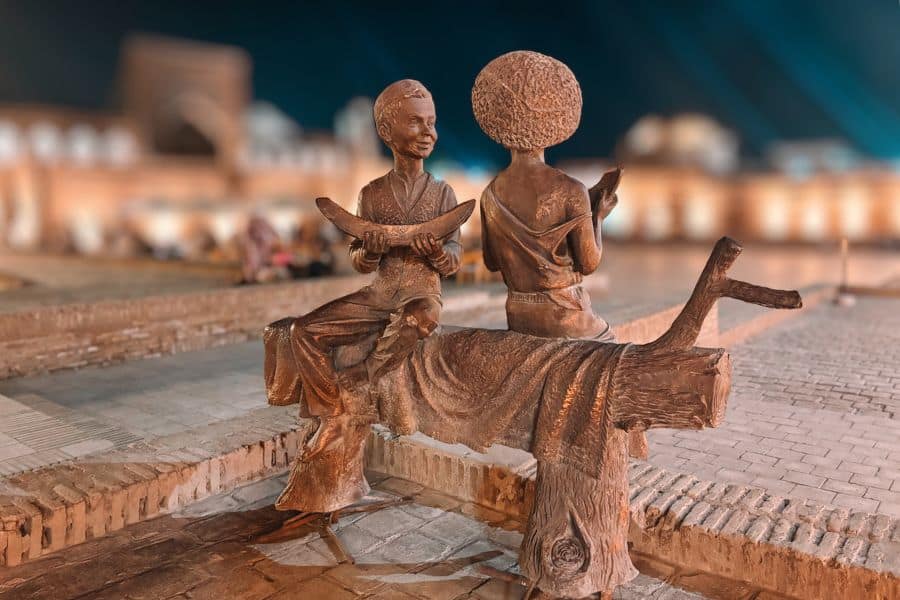 bronze statue of khiva children sitting on a bench at night in itchan kala with illuminated historic buildings in background