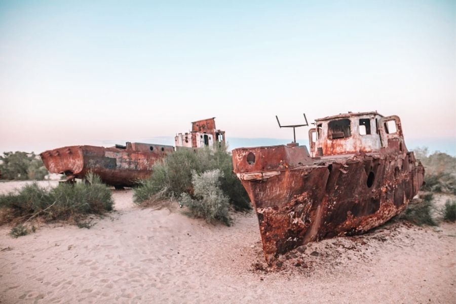 rusted ships lying in the sands of the moynaq ship cemetery in uzbekistan, once part of the aral sea, surrounded by dry desert shrubs under a soft pastel evening sky
