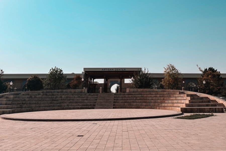 wide view of the artisan center in shahrisabz with stone steps, circular open plaza, and a shaded entrance pavilion framed by trees under a clear blue sky