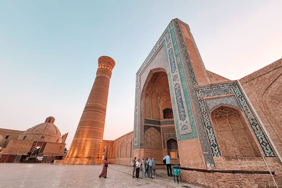 poi kalyan complex in bukhara at sunset with people walking near the illuminated minaret and turquoise-tiled madrasa under a clear sky