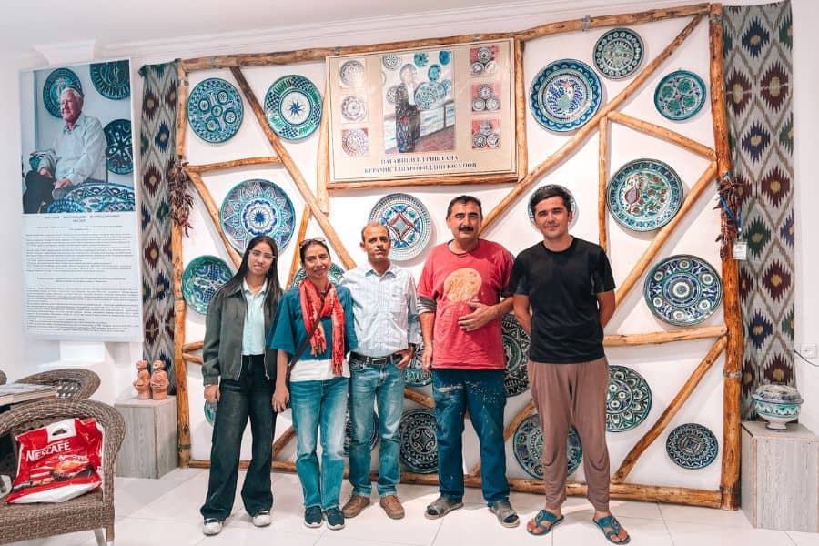 our family posing with uzbek potters at sharofiddin yusupov’s ceramics workshop in rishton, run by his son and grandson, surrounded by vibrant hand-painted blue pottery inside the international ceramics centre