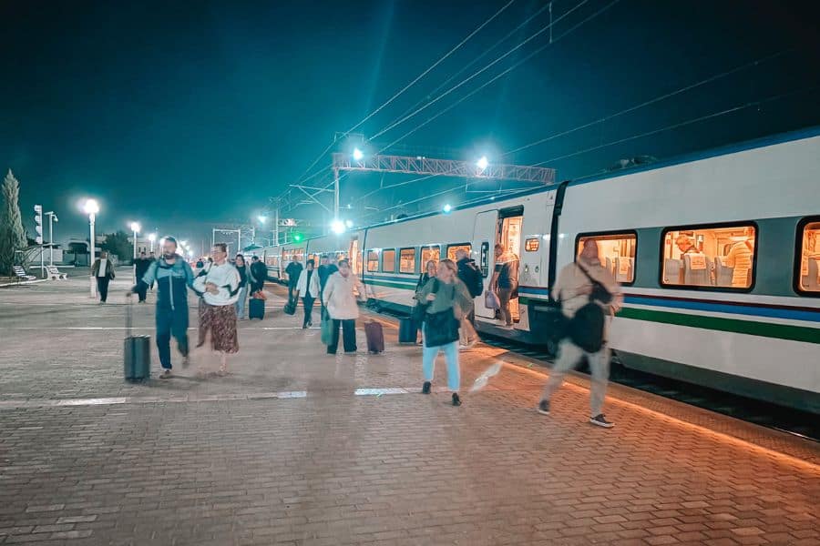 passengers boarding the afrosiyab high speed train at samarkand railway station at night, with illuminated train coaches and people walking along the platform with luggage