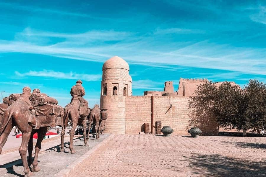 camel caravan sculpture outside itchan kala fortress walls in khiva uzbekistan, with mudbrick minaret and historic city walls under blue sky