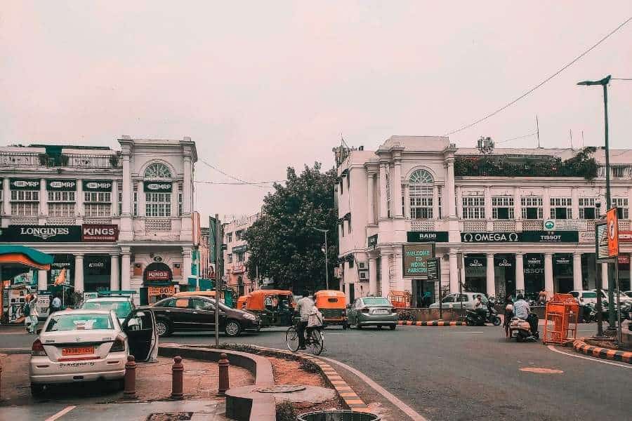 a busy street scene at connaught place, delhi, with colonial-era white buildings housing brand stores like omega and tissot, auto-rickshaws, cars, and cyclists navigating a traffic circle under an overcast sky