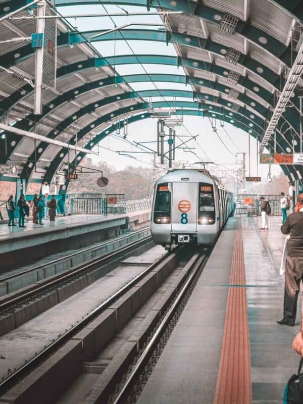 a delhi metro train arriving at an elevated station with a curved roof, passengers waiting on the platform, and overhead electric wires stretching into the distance