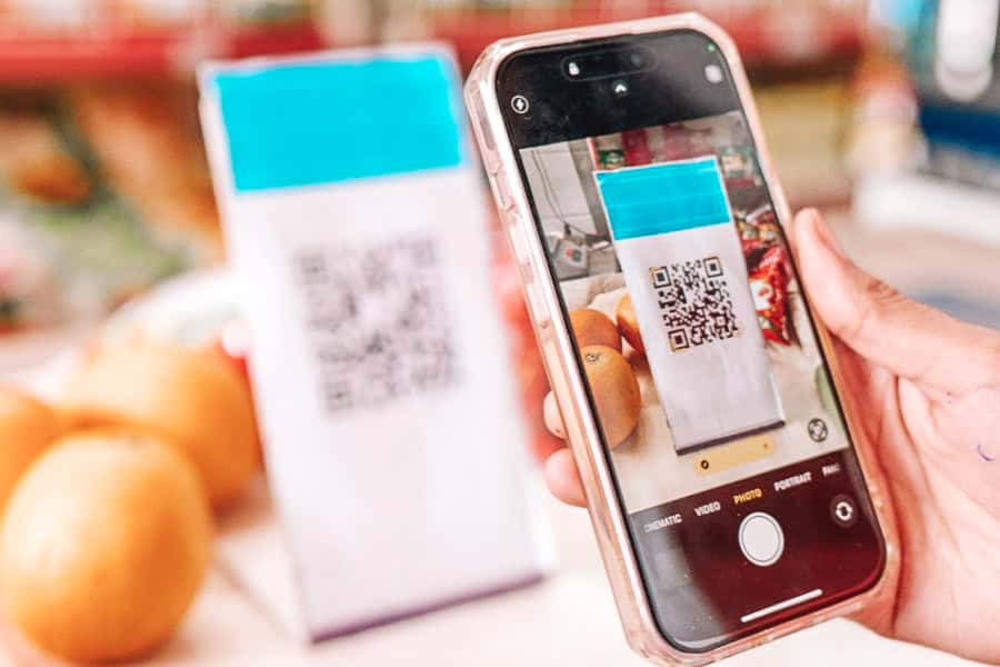 a person scanning a qr code using a smartphone for digital payment at a local fruit shop, with oranges in the background and the blurred code stand visible behind the phone