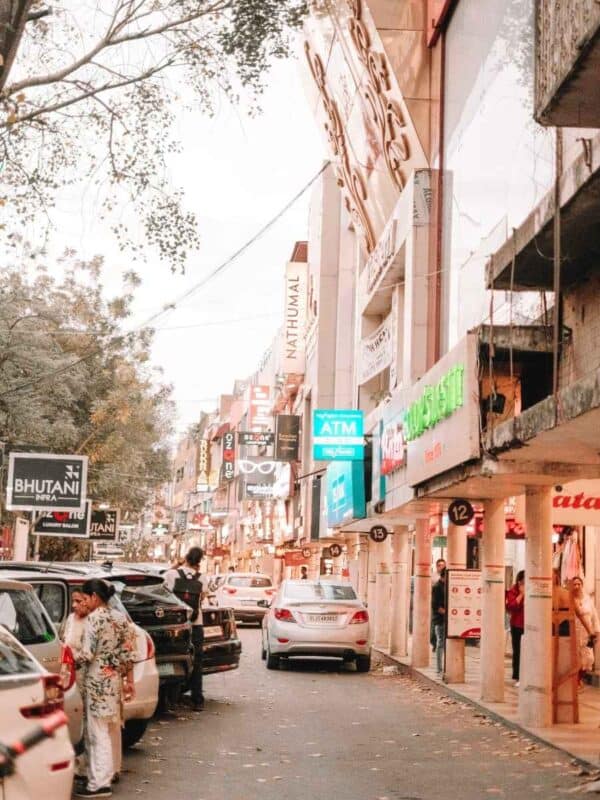 a lively evening scene at gk1 m block market in delhi, showing shoppers, parked cars, and glowing storefronts along a narrow street
