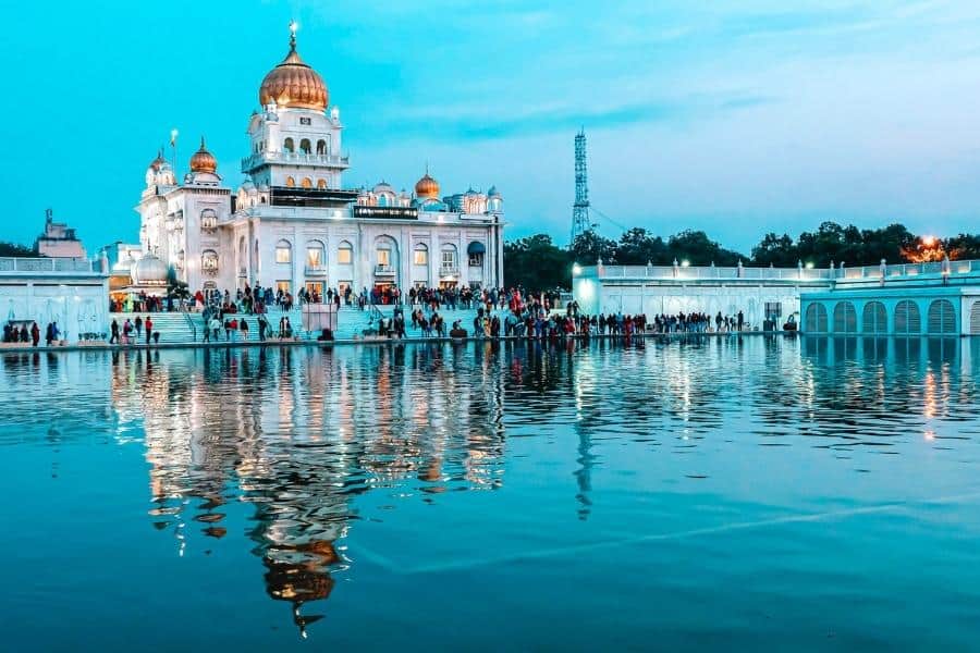 evening view of gurudwara bangla sahib with golden domes glowing and reflecting in the sacred pond, surrounded by devotees and city skyline in the backdrop