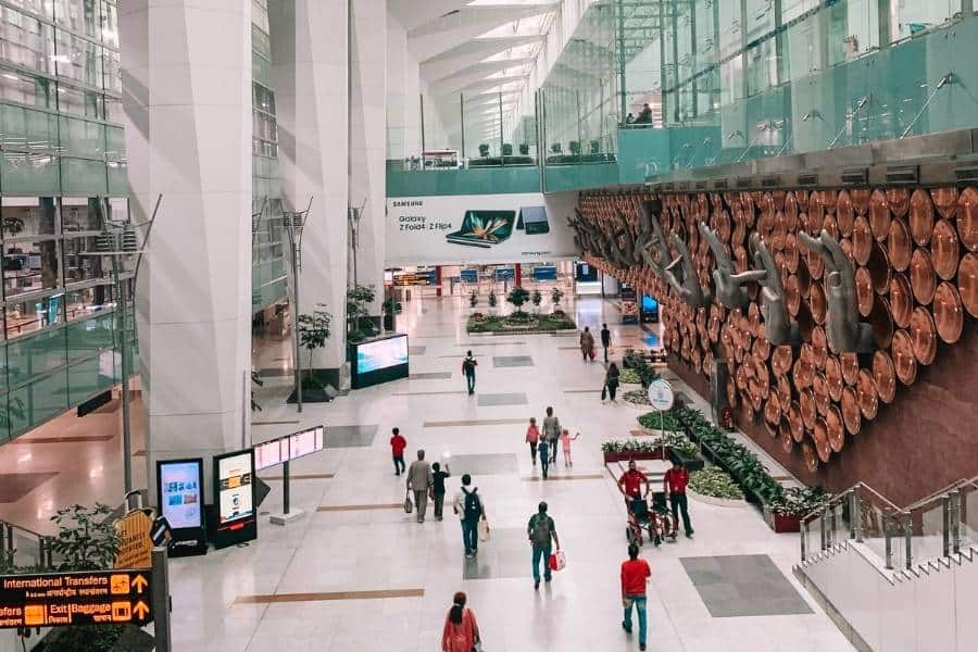 inside view of delhi’s indira gandhi international airport terminal, with passengers walking across the spacious, modern concourse, and the iconic hand mudra wall installation displayed prominently on the right side
