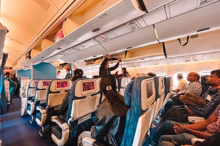 inside an uzbekistan airways flight from tashkent to delhi showing economy class seating, passengers boarding, open overhead bins, and seatback screens along the aisle